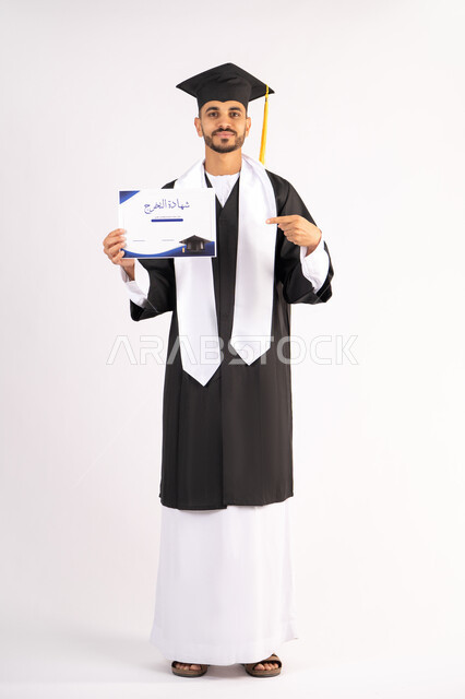 Pointing the index finger at the certificate, achieving the dream of success, university occasion and celebration, celebrating the completion of university studies, the concept of diligence and excellence, portrait of a smiling young Arab Gulf Emirati graduate wearing a graduation gown looking at the camera with an expression of happiness, full body, white background