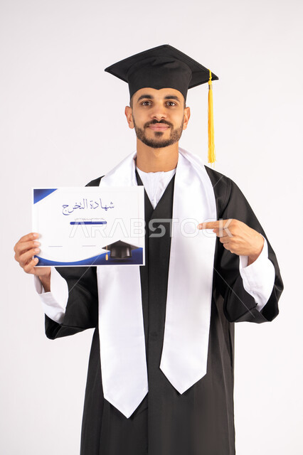 الاشارة باصبع السبابة إلى الشهادة، الوصول لحلم النجاح، مناسبة واحتفال جامعي، الاحتفال بإنهاء الدراسة الجامعية، مفهوم الاجتهاد والتفوق، بورتريه لشاب خريج عربي خليجي اماراتي مبتسم يرتدي عباءة التخرج ينظر الى الكاميرا بتعابير السعادة ،  خلفية بيضاء
