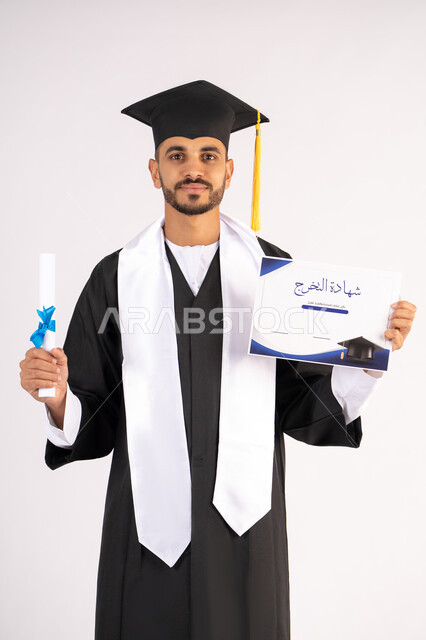 حمل الشهادة بتعابير الفروح والسرور، الشعور بالفخر والثقة بالنفس، الاحتفال بالنجاح وانهاء الدراسة الجامعية، مفهوم التفوق والاجتهاد، بورتريه لخريج عربي خليجي اماراتي يرتدي قبعة وثوب التخرج يقف باستقامة بايماءات السعادة، خلفية بيضاء