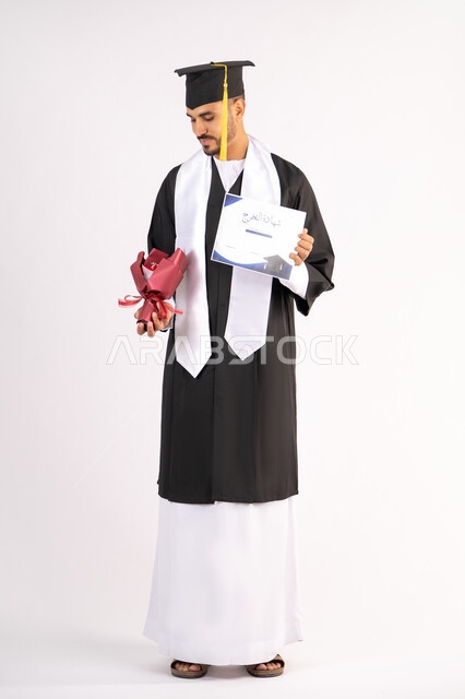 الشعور بالفخر والثقة بالنفس، استلام الشهادة بتعابير الفروح والسرور، الاحتفال بالنجاح وانهاء الدراسة الجامعية، مفهوم التفوق والاجتهاد، بورتريه لخريج عربي خليجي اماراتي يرتدي قبعة وثوب التخرج يقوم بأخذ باقة الورد بايماءات السعادة، خلفية بيضاء