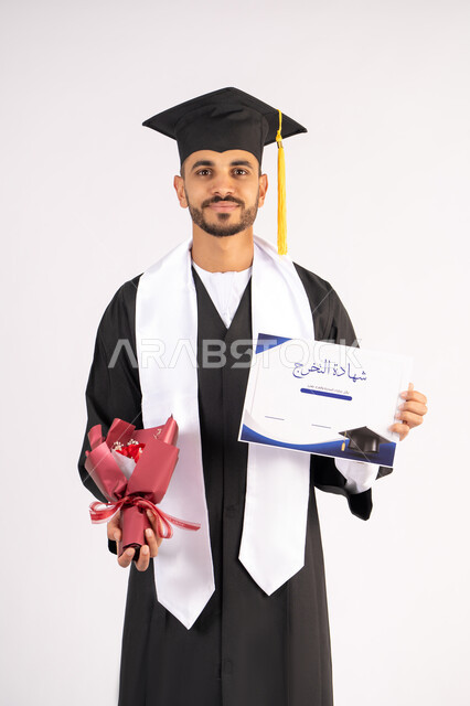 استلام الشهادة بتعابير الفرح، الشعور بالفخر والثقة بالنفس، الاحتفال بالنجاح وانهاء الدراسة الجامعية، مفهوم التفوق والاجتهاد، بورتريه لشاب خريج عربي خليجي اماراتي يرتدي قبعة وثوب التخرج يمسك بيده باقة الورد بايماءات السعادة، خلفية بيضاء