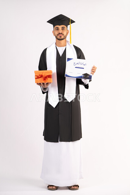 الحصول على الشهادة بتعابير الفرح، الشعور بالفخر والثقة بالنفس، الاحتفال بالنجاح وانهاء الدراسة الجامعية، مفهوم التفوق والاجتهاد، بورتريه لشاب خريج عربي خليجي اماراتي يرتدي قبعة وثوب التخرج يمسك بيده هدية بايماءات السعادة، الجسم بالطول الكامل، خلفية بيضاء