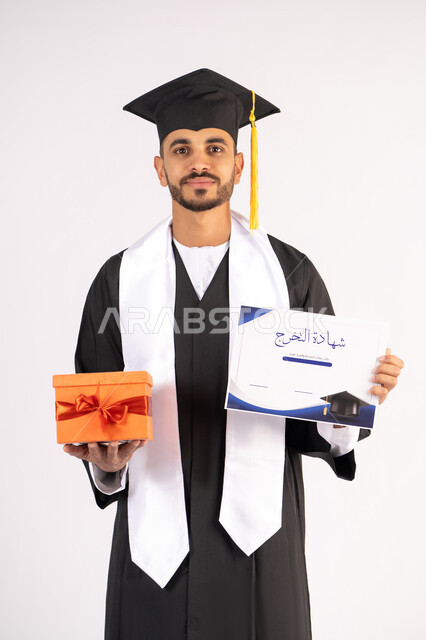 Obtaining a certificate with expressions of joy, feeling proud and confident, celebrating success and completing university studies, the concept of excellence and diligence, portrait of a young Arab Gulf Emirati graduate wearing a graduation cap and gown holding a gift in his hand with gestures of happiness , white background