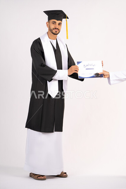 اخذ الشهادة بتعابير الفرح، الشعور بالفخر والثقة بالنفس، الاحتفال بالنجاح وانهاء الدراسة الجامعية، مفهوم التفوق والاجتهاد، بورتريه لشاب خريج عربي خليجي اماراتي يرتدي قبعة وثوب التخرج ينظر الى الكاميرا بايماءات السعادة، الجسم بالطول الكامل، خلفية بيضاء