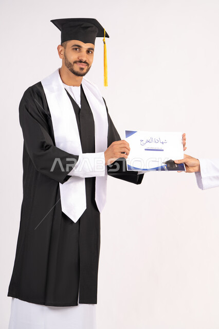 اخذ الشهادة بتعابير الفرح، الشعور بالفخر والثقة بالنفس، الاحتفال بالنجاح وانهاء الدراسة الجامعية، مفهوم التفوق والاجتهاد، بورتريه لشاب خريج عربي خليجي اماراتي يرتدي قبعة وثوب التخرج ينظر الى الكاميرا بايماءات السعادة، خلفية بيضاء