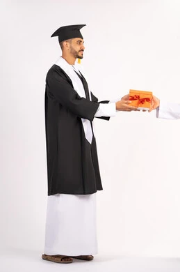 تقديم الهدايا بتعابير الفرح، الشعور بالفخر والثقة بالنفس، الاحتفال بالنجاح وانهاء الدراسة الجامعية، مفهوم التفوق والاجتهاد، بورتريه لشاب خريج عربي خليجي اماراتي يرتدي قبعة وثوب التخرج يقوم بأخذ الهدية بايماءات السعادة، الجسم بالطول الكامل، خلفية بيضاء