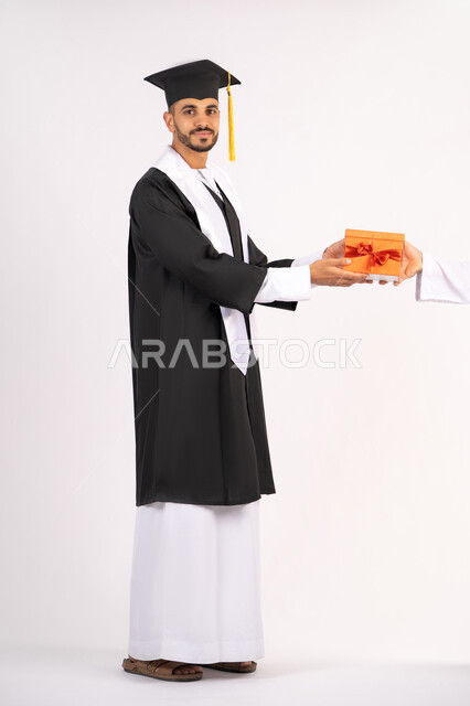 تقديم الهدايا بتعابير الفرح، الشعور بالفخر والثقة بالنفس، الاحتفال بالنجاح وانهاء الدراسة الجامعية، مفهوم التفوق والاجتهاد، بورتريه لشاب خريج عربي خليجي اماراتي يرتدي قبعة وثوب التخرج يقوم بأخذ الهدية بايماءات السعادة، الجسم بالطول الكامل، خلفية بيضاء