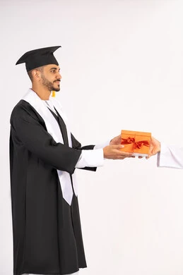 تقديم الهدايا بتعابير الفرح، الشعور بالفخر والثقة بالنفس، الاحتفال بالنجاح وانهاء الدراسة الجامعية، مفهوم التفوق والاجتهاد، بورتريه لشاب خريج عربي خليجي اماراتي يرتدي قبعة وثوب التخرج يقوم بأخذ الهدية بايماءات السعادة ، خلفية بيضاء