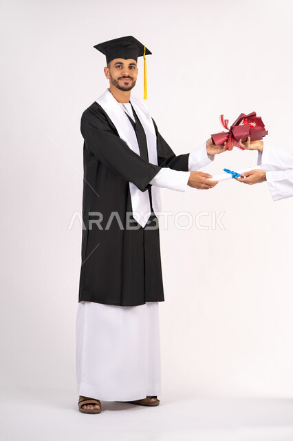 النظر الى الكاميرا بتعابير السرور والثقة بالنفس، استلام الشهادة بايماءات الفروح والسرور، الاحتفال بالنجاح وانهاء الدراسة الجامعية، مفهوم التفوق والاجتهاد، بورتريه لخريج عربي خليجي اماراتي يرتدي قبعة وثوب التخرج يقوم بأخذ باقة الورد، الجسم بالطول الكامل، خلفية بيضاء