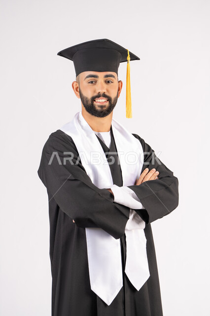 الجد والنجاح والاجتهاد، التقاط صور شخصية بثياب التخرج، الاحتفال بإنهاء المرحلة الجامعية، بورتريه لشاب خريج عربي خليجي اماراتي مبتسم يرتدي القبعة وعباءة التخرج يقف بأيدي متقاطعة ينظر الى الكاميرا بتعابير الثقة بالنفس، خلفية بيضاء