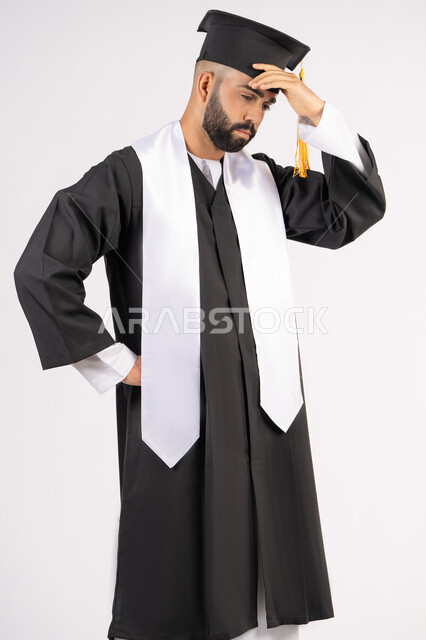 الارهاق والتعب بعد الاجتهاد والمثابرة، الانتهاء من مرحلة الدراسة الجامعية بعد عناء، بورتريه لشاب خريج عربي خليجي اماراتي يرتدي القبعة وزي التخرج يضع يده على رأسه بتعابير اليأس،  انتظار النتائج بفارغ الصبر، خلفية بيضاء