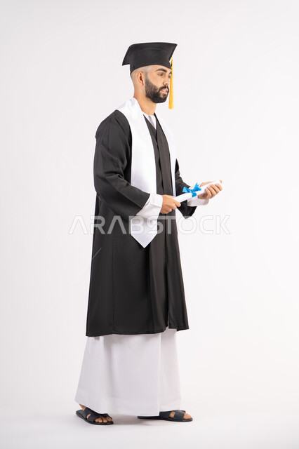 حمل الشهادة بتعابير الفروح والسرور، الشعور بالفخر والثقة بالنفس، الاحتفال بالنجاح وانهاء الدراسة الجامعية، مفهوم التفوق والاجتهاد، بورتريه لخريج عربي خليجي اماراتي يرتدي قبعة وثوب التخرج يقف باستقامة بايماءات السعادة، الجسم بالطول الكامل، خلفية بيضاء