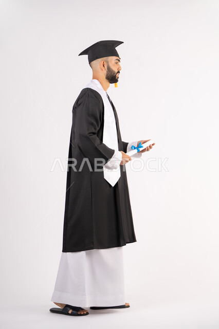 Achieving the dream of success, expressions of happiness and pleasure, university occasion and celebration, celebration of completing university studies, concept of diligence and excellence, portrait of a smiling young Arab Gulf Emirati graduate wearing a graduation gown holding a certificate in his hand, full body, white background