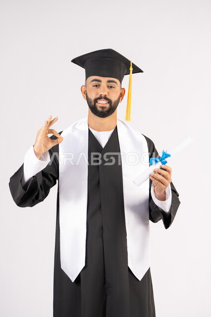 A mark of quality, excellence and distinction, celebrating the achievement of dreams, celebrating the completion of university, a portrait of a young Arab Gulf Emirati graduate wearing a graduation cap and gown, holding a certificate in his hand, looking at the camera with happy gestures, white background