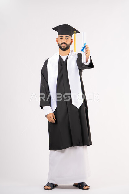 حمل الشهادة بتعابير الفرح والسرور، الشعور بالفخر والثقة بالنفس، الاحتفال بالنجاح وانهاء الدراسة الجامعية، مفهوم التفوق والاجتهاد، بورتريه لخريج عربي خليجي اماراتي يرتدي قبعة وثوب التخرج يقف باستقامة بايماءات السعادة، الجسم بالطول الكامل، خلفية بيضاء