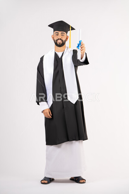 الوصول لحلم النجاح، تعابير السعادة والسرور، مناسبة واحتفال جامعي، الاحتفال بإنهاء الدراسة الجامعية، مفهوم الاجتهاد والتفوق، بورتريه لشاب خريج عربي خليجي اماراتي مبتسم يرتدي عباءة التخرج يمسك بيده الشهادة، الجسم بالطول الكامل، خلفية بيضاء