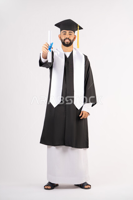 Achieving the dream of success, expressions of happiness and pleasure, university occasion and celebration, celebration of completing university studies, concept of diligence and excellence, portrait of a smiling young Arab Gulf Emirati graduate wearing a graduation gown holding a certificate in his hand, full body, white background