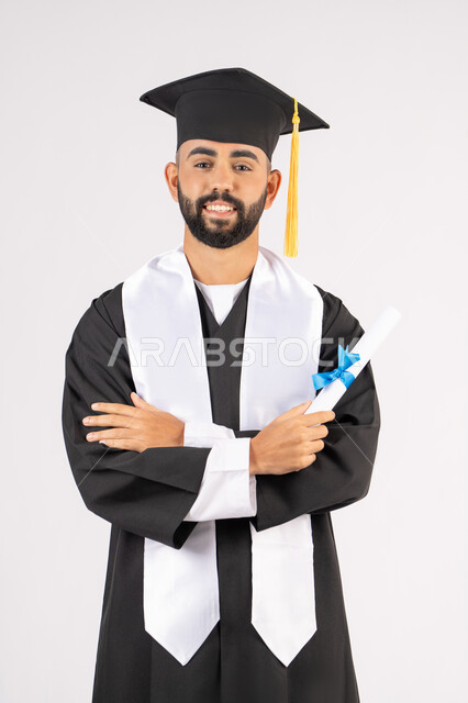 التقاط صور شخصية بثياب التخرج، الاحتفال بإنهاء المرحلة الجامعية، بورتريه لشاب خريج عربي خليجي اماراتي مبتسم يرتدي القبعة وعباءة التخرج يقف بأيدي متقاطعة ينظر الى الكاميرا بتعابير الثقة بالنفس، مفهوم التفوق والنجاح والاجتهاد، خلفية بيضاء
