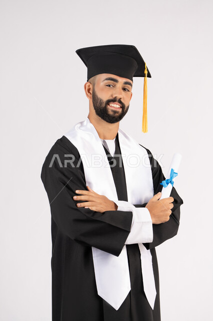 التقاط صور شخصية بثياب التخرج، الاحتفال بإنهاء المرحلة الجامعية، بورتريه لشاب خريج عربي خليجي اماراتي مبتسم يرتدي القبعة وعباءة التخرج يقف بأيدي متقاطعة ينظر الى الكاميرا بتعابير الثقة بالنفس، مفهوم التفوق والنجاح والاجتهاد، خلفية بيضاء