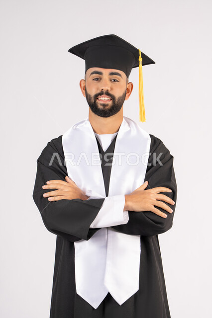 التقاط صور شخصية بثياب التخرج، الاحتفال بإنهاء المرحلة الجامعية، بورتريه لشاب خريج عربي خليجي اماراتي مبتسم يرتدي القبعة وعباءة التخرج يقف بأيدي متقاطعة ينظر الى الكاميرا بتعابير الثقة بالنفس، مفهوم التفوق والنجاح والاجتهاد، خلفية بيضاء