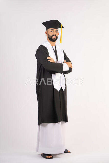 The concept of excellence, success and diligence, taking selfies in graduation gowns, celebrating the completion of university, portrait of a smiling young Arab Gulf Emirati graduate wearing a graduation cap and gown, standing with crossed hands looking at the camera with an expression of self-confidence, full body, white background