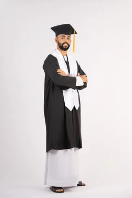 مفهوم التفوق والنجاح والاجتهاد، التقاط صور شخصية بثياب التخرج، الاحتفال بإنهاء المرحلة الجامعية، بورتريه لشاب خريج عربي خليجي اماراتي مبتسم يرتدي القبعة وعباءة التخرج يقف بأيدي متقاطعة ينظر الى الكاميرا بتعابير الثقة بالنفس، الجسم بالطول الكامل، خلفية بيضاء