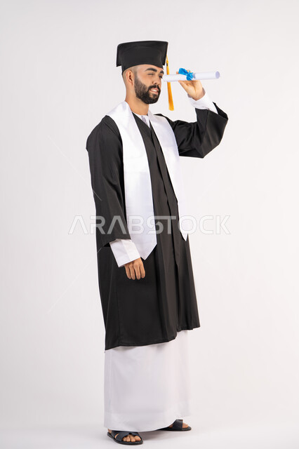 الفخر والفرح بالوصول لحلم التخرج, التطلع إلى المستقبل بعد التخرج، بورتريه من الجانب لشاب خريج عربي سعودي خليجي يرتدي ثياب التخرج ينظر من خلال شهادة التفوق، الاحتفال بانتهاء المرحلة الجامعية، الجسم بالطول الكامل، خلفية بيضاء