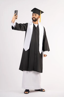 توثيق اللحظات السعيدة، استخدام التقنية الحديثة، الاحتفال بإنهاء الدراسة الجامعية، بورتريه لشاب خريج عربي خليجي اماراتي يرتدي عباءه وقبعة التخرج  يقوم بالتقاط صورة سيلفي تذكارية عبر الهاتف المحمول، الجسم بالطول الكامل، خلفية بيضاء