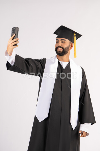 الاحتفاظ بالذكريات السعيدة، استخدام التقنية الحديثة، الاحتفال بإنهاء الدراسة الجامعية، بورتريه لشاب خريج عربي خليجي اماراتي يرتدي عباءه وقبعة التخرج  يقوم بالتقاط صورة سيلفي تذكارية عبر الهاتف المحمول، خلفية بيضاء