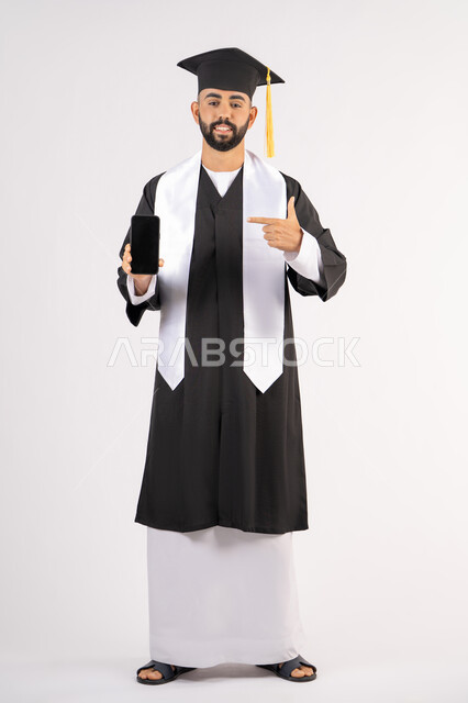 Blank black screen on mobile phone, online education and study applications, portrait of a smiling young Arab Gulf Emirati graduate wearing a cap and graduation gown with a happy expression, celebrating the end of the university stage, full body, white background