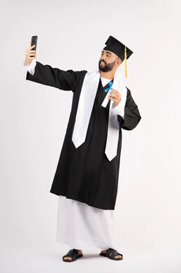 توثيق اللحظات السعيدة، استخدام التقنية الحديثة، الاحتفال بإنهاء الدراسة الجامعية، بورتريه لشاب خريج عربي خليجي اماراتي يرتدي عباءه وقبعة التخرج  يقوم بالتقاط صورة سيلفي تذكارية عبر الهاتف المحمول، الجسم بالطول الكامل، خلفية بيضاء