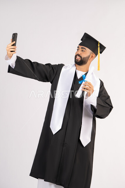 الاحتفاظ بالذكريات السعيدة، استخدام التقنية الحديثة، الاحتفال بإنهاء الدراسة الجامعية، بورتريه لشاب خريج عربي خليجي اماراتي يرتدي عباءه وقبعة التخرج  يقوم بالتقاط صورة سيلفي تذكارية عبر الهاتف المحمول، خلفية بيضاء