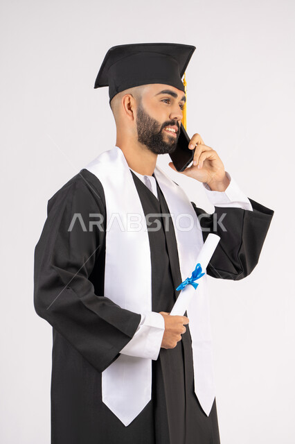 التواصل مع الأهل والأصدقاء، مفهوم الاجتهاد والتفوق، الاحتفال بإنهاء الدراسة الجامعية، استخدام التقنية الحديثة، بورتريه مقرب لشاب خريج عربي خليجي اماراتي يرتدي عباءه وقبعة التخرج يقوم بإجراء مكالمة عبر الهاتف المحمول، خلفية بيضاء