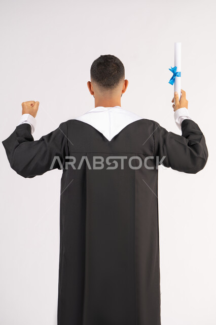الحصول على الشهادة، التعليم في الامارات العربية المتحدة، الشعور بالفخر والفرح بالوصول لحلم التخرج, بورتريه من الخلف لشاب خريج عربي خليجي اماراتي يرتدي عباءة يرفع وثيقة التخرج، الاحتفال بإنهاء المرحلة الجامعية، خلفية بيضاء