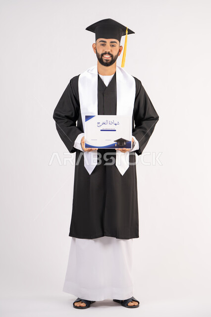 الوصول لحلم النجاح، تعابير السعادة والسرور، مناسبة واحتفال جامعي، الاحتفال بإنهاء الدراسة الجامعية، مفهوم الاجتهاد والتفوق، بورتريه لشاب خريج عربي خليجي اماراتي مبتسم يرتدي عباءة التخرج يمسك بيده الشهادة، الجسم بالطول الكامل، خلفية بيضاء