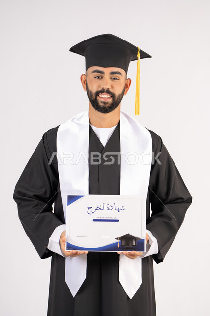 Holding a certificate with expressions of joy and happiness, feeling proud and self-confident, celebrating success and completing university studies, the concept of excellence and diligence, a portrait of an Arab Gulf Emirati graduate wearing a graduation cap and gown, standing straight with gestures of happiness, white background