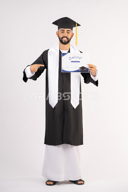 الاشارة باصبع السبابة إلى الشهادة، الوصول لحلم النجاح، مناسبة واحتفال جامعي، الاحتفال بإنهاء الدراسة الجامعية، مفهوم الاجتهاد والتفوق، بورتريه لشاب خريج عربي خليجي اماراتي مبتسم يرتدي عباءة التخرج ينظر الى الكاميرا بتعابير السعادة ، الجسم بالطول الكامل، خلفية بيضاء