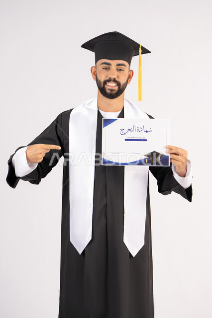 الاشارة باصبع السبابة إلى الشهادة، الوصول لحلم النجاح، مناسبة واحتفال جامعي، الاحتفال بإنهاء الدراسة الجامعية، مفهوم الاجتهاد والتفوق، بورتريه لشاب خريج عربي خليجي اماراتي مبتسم يرتدي عباءة التخرج ينظر الى الكاميرا بتعابير السعادة ، خلفية بيضاء