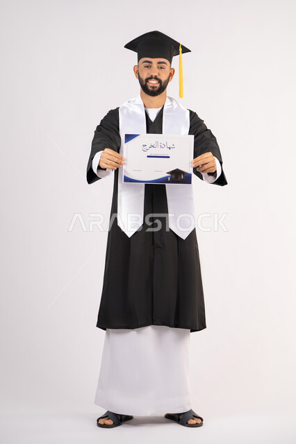 الوصول لحلم النجاح، تعابير السعادة والسرور، مناسبة واحتفال جامعي، الاحتفال بإنهاء الدراسة الجامعية، مفهوم الاجتهاد والتفوق، بورتريه لشاب خريج عربي خليجي اماراتي مبتسم يرتدي عباءة التخرج يمسك بيده الشهادة، الجسم بالطول الكامل، خلفية بيضاء
