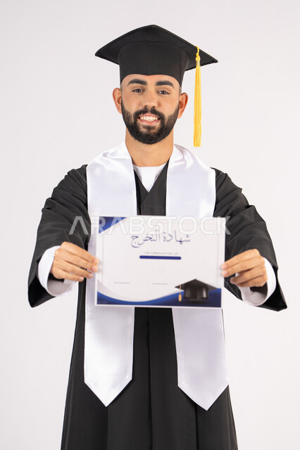 جودة التعليم في الامارات، الوصول لحلم النجاح، تعابير السعادة والسرور، مناسبة النجاح في الجامعة، الاحتفال بإنهاء الدراسة الجامعية، مفهوم الاجتهاد والتفوق، بورتريه لشاب خريج عربي خليجي اماراتي مبتسم يرتدي عباءة التخرج يمسك بيده الشهادة، خلفية بيضاء