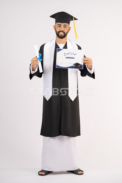 Expressions of happiness and joy, achieving the dream of success, university occasion and celebration, celebration of completing university studies, concept of diligence and excellence, portrait of a smiling young Arab Gulf Emirati graduate wearing a graduation gown holding a certificate in his hand, full body, white background