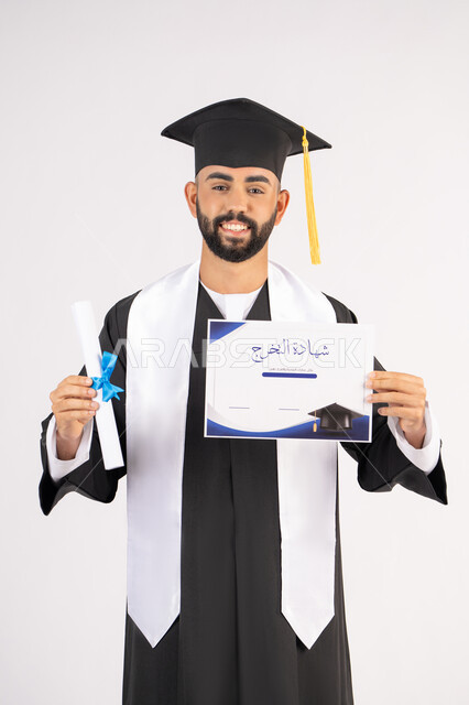 جودة التعليم في الامارات، الوصول لحلم النجاح، تعابير السعادة والسرور، مناسبة النجاح في الجامعة، الاحتفال بإنهاء الدراسة الجامعية، مفهوم الاجتهاد والتفوق، بورتريه لشاب خريج عربي خليجي اماراتي مبتسم يرتدي عباءة التخرج يمسك بيده الشهادة، خلفية بيضاء