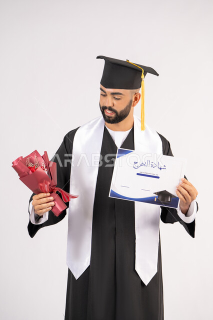 إيماءات السعادة والسرور بالوصول لحلم النجاح، مناسبة واحتفال جامعي، الاحتفال بإنهاء الدراسة الجامعية، مفهوم الاجتهاد والتفوق، بورتريه لشاب خريج عربي خليجي اماراتي مبتسم يرتدي عباءة التخرج يمسك بيده باقة ورد والشهادة، خلفية بيضاء