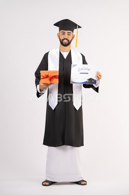 الحصول على وثيقة التخرج، تقديم الهدايا في المناسبات، الاحتفال بإنهاء الدراسة الجامعية، إيماءات السعادة بالوصول لحلم النجاح، بورتريه لشاب خريج عربي خليجي اماراتي يرتدي زي التخرج والقبعة يحمل بيده الشهادة وصندوق الهدية، الجسم بالطول الكامل، خلفية بيضاء