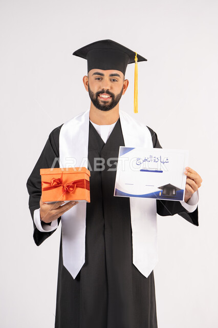 Obtaining a graduation certificate, giving gifts on occasions, celebrating the completion of university studies, gestures of happiness for reaching the dream of success, portrait of a young Arab Gulf Emirati graduate wearing graduation attire and cap, holding a certificate and gift box, white background