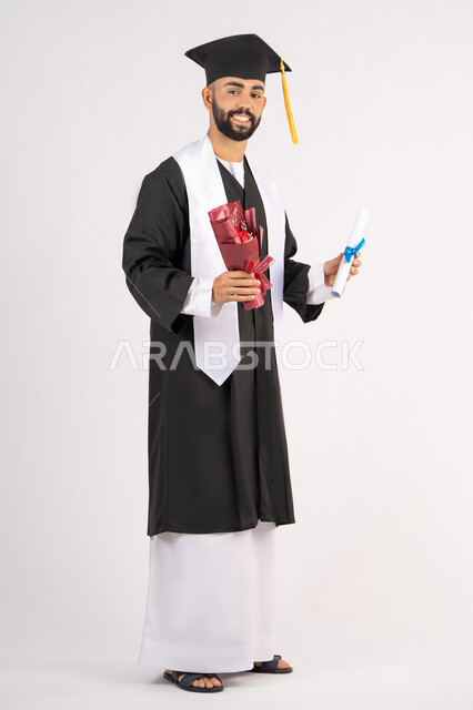 Expressions of happiness and joy at achieving the dream of success, a university occasion and celebration, celebrating the completion of university studies, the concept of diligence and excellence, a portrait of a smiling young Arab Gulf Emirati graduate wearing a graduation gown, holding a bouquet of flowers and a certificate, full body, white background