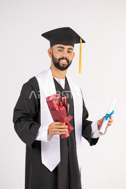 إيماءات السعادة والسرور بالوصول لحلم النجاح، مناسبة واحتفال جامعي، الاحتفال بإنهاء الدراسة الجامعية، مفهوم الاجتهاد والتفوق، بورتريه لشاب خريج عربي خليجي اماراتي مبتسم يرتدي عباءة التخرج يمسك بيده باقة ورد والشهادة، خلفية بيضاء