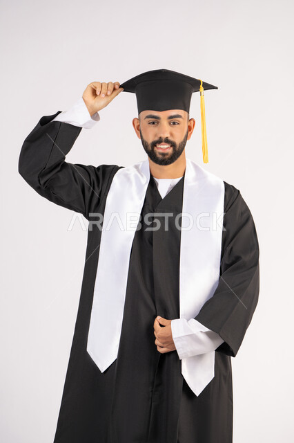 الاجتهاد والتفوق والنجاح، الاحتفال بإنهاء المرحلة الجامعية، تعابير السرور والسعادة، التقاط صور شخصية بثياب التخرج، بورتريه لشاب خريج عربي خليجي اماراتي مبتسم يرتدي عباءة التخرج يمسك بيده القبعة ينظر الى الكاميرا بإيماءات البهجة، خلفية بيضاء