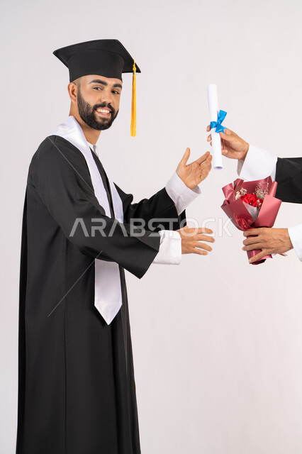 الشعور بالفخر والثقة بالنفس، استلام الشهادة بتعابير الفروح والسرور، مفهوم التفوق والاجتهاد، بورتريه من الجانب لخريج عربي خليجي اماراتي يرتدي قبعة وثوب التخرج يقوم بأخذ باقة الورد بايماءات السعادة، الاحتفال بالنجاح وانهاء الدراسة الجامعية، خلفية بيضاء