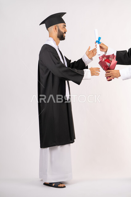 استلام الشهادة بتعابير الفروح والسرور، مفهوم التفوق والاجتهاد، بورتريه من الجانب لخريج عربي خليجي اماراتي يرتدي قبعة وثوب التخرج يقوم بأخذ باقة الورد بايماءات السعادة، الاحتفال بالنجاح وانهاء الدراسة الجامعية، صورة الجسم بالطول الكامل، خلفية بيضاء
